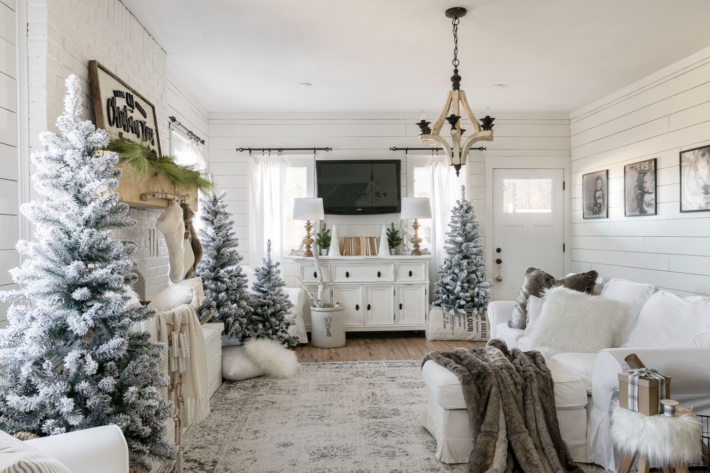 The white Christmas home living room decorated with several snow-crusted pine trees and many pillows and blankets in a neutral color scheme.