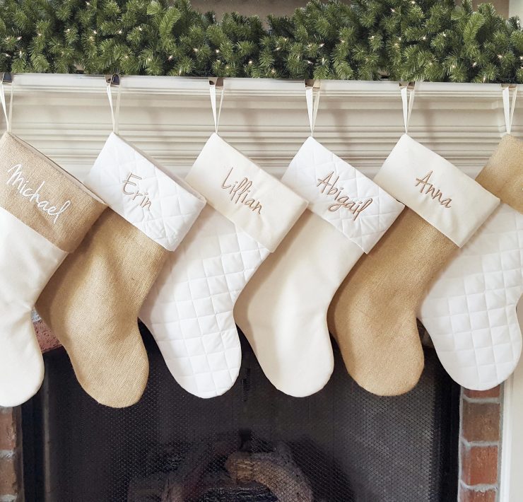 Six personalized stockings in varying neutral shades hanging on a fireplace mantel.
