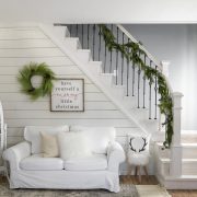 Small, white couch beneath a garland-adorned staircase and white shiplap wall, perfect for the neutral Christmas look.