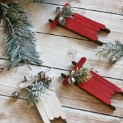 Three DIY ornaments made with painted popsicle sticks, spruce tips and mini silver bells made to look like sleighs.