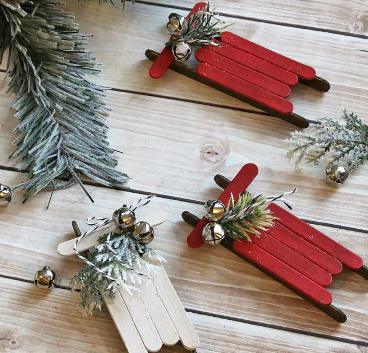 Three DIY ornaments made with painted popsicle sticks, spruce tips and mini silver bells made to look like sleighs.