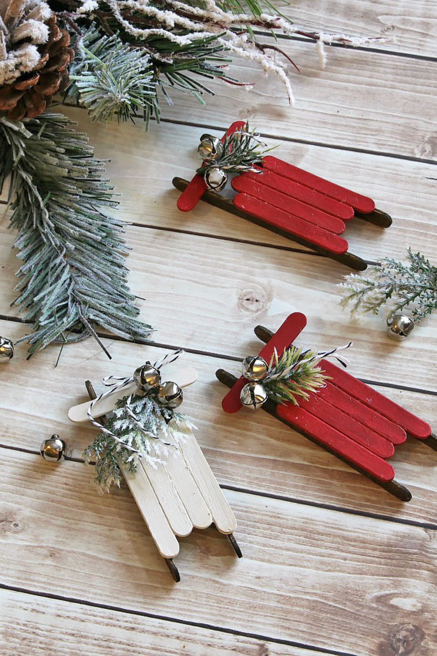 Three DIY ornaments made with painted popsicle sticks, spruce tips and mini silver bells made to look like sleighs.