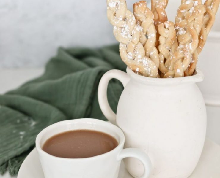 Braided sugar cookie sticks in a milk jug beside a cup of hot cocoa