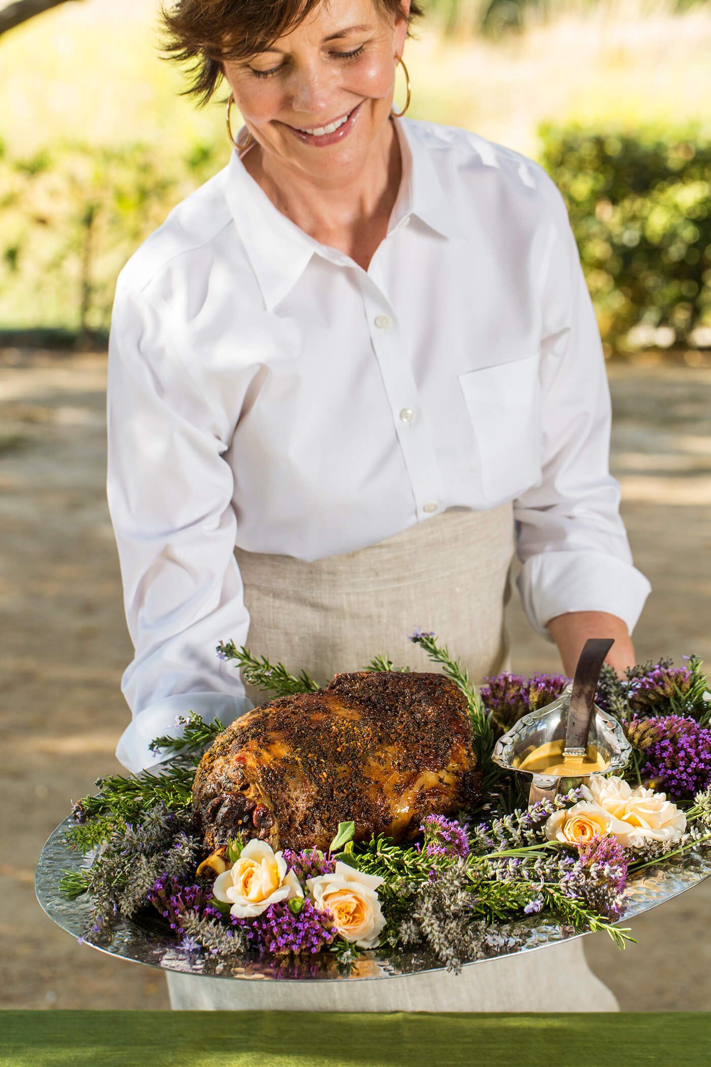 A woman is holding a platter covered in the slow roasted leg of lamb framed by freshly cut spring roses in the colors of purple and light orange