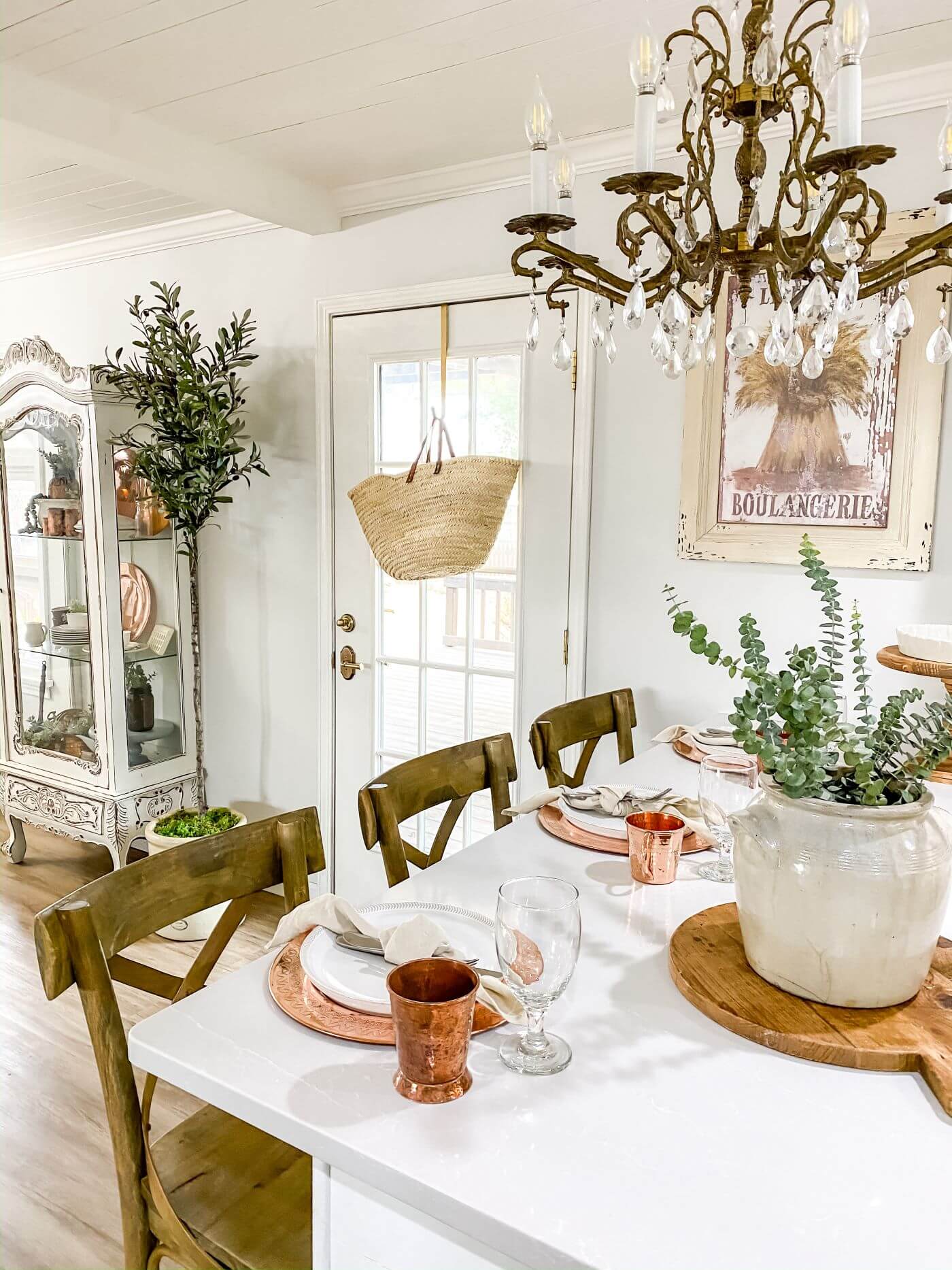 A white farmhouse table with an antique crystal chandelier above. The table settings are made from copper colored material and a white ceramic piece houses fresh pops of green from the garden in the table’s centerpiece