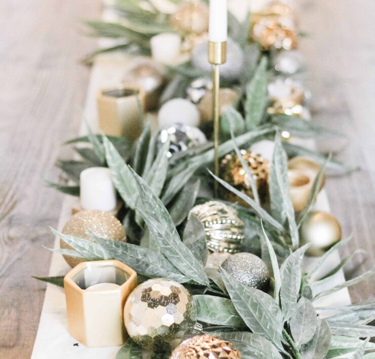 close up of center tablescape full of fresh green garland, gold ornaments, and simple white candles with a white table runner