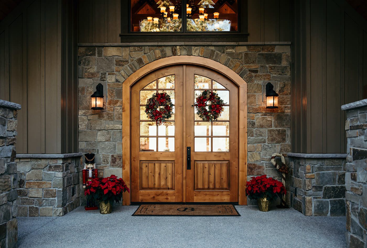 Wreaths hang from wood double doors framed by glass sconces and red flowers.